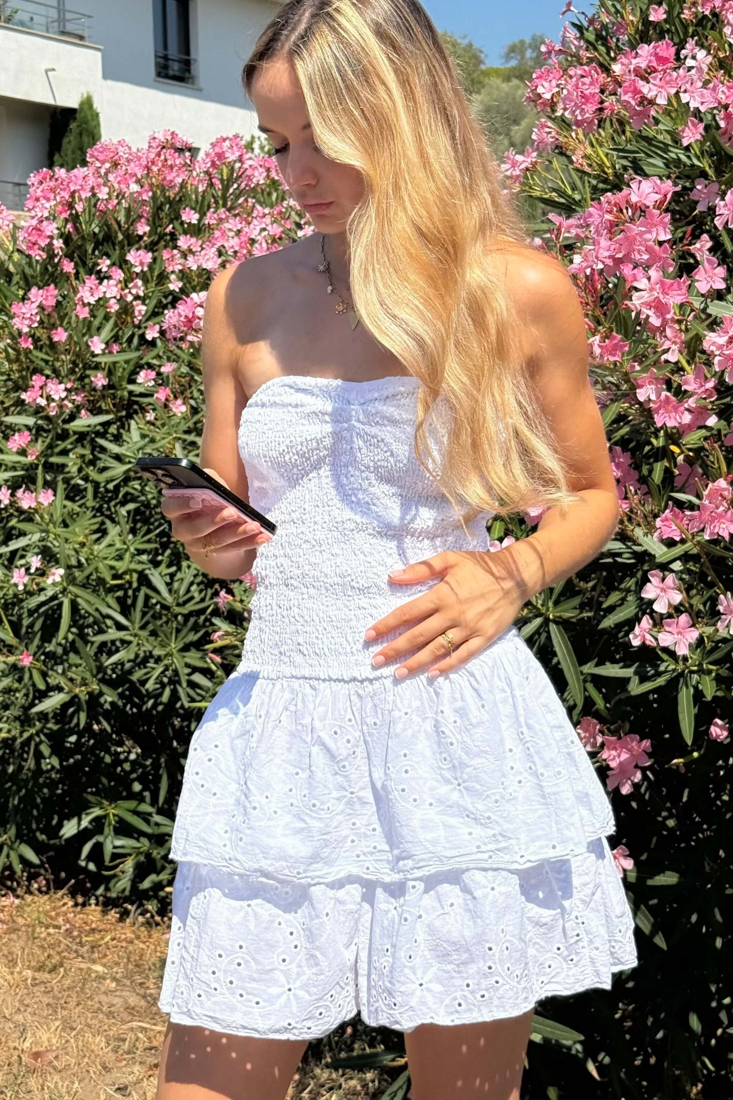 robe bustier blanche femme en coton avec détails broderie anglaise et haut smocké élastique. Mise en avant de la texture ajourée et du tissu léger. Parfait pour un look d’été chic et confortable.