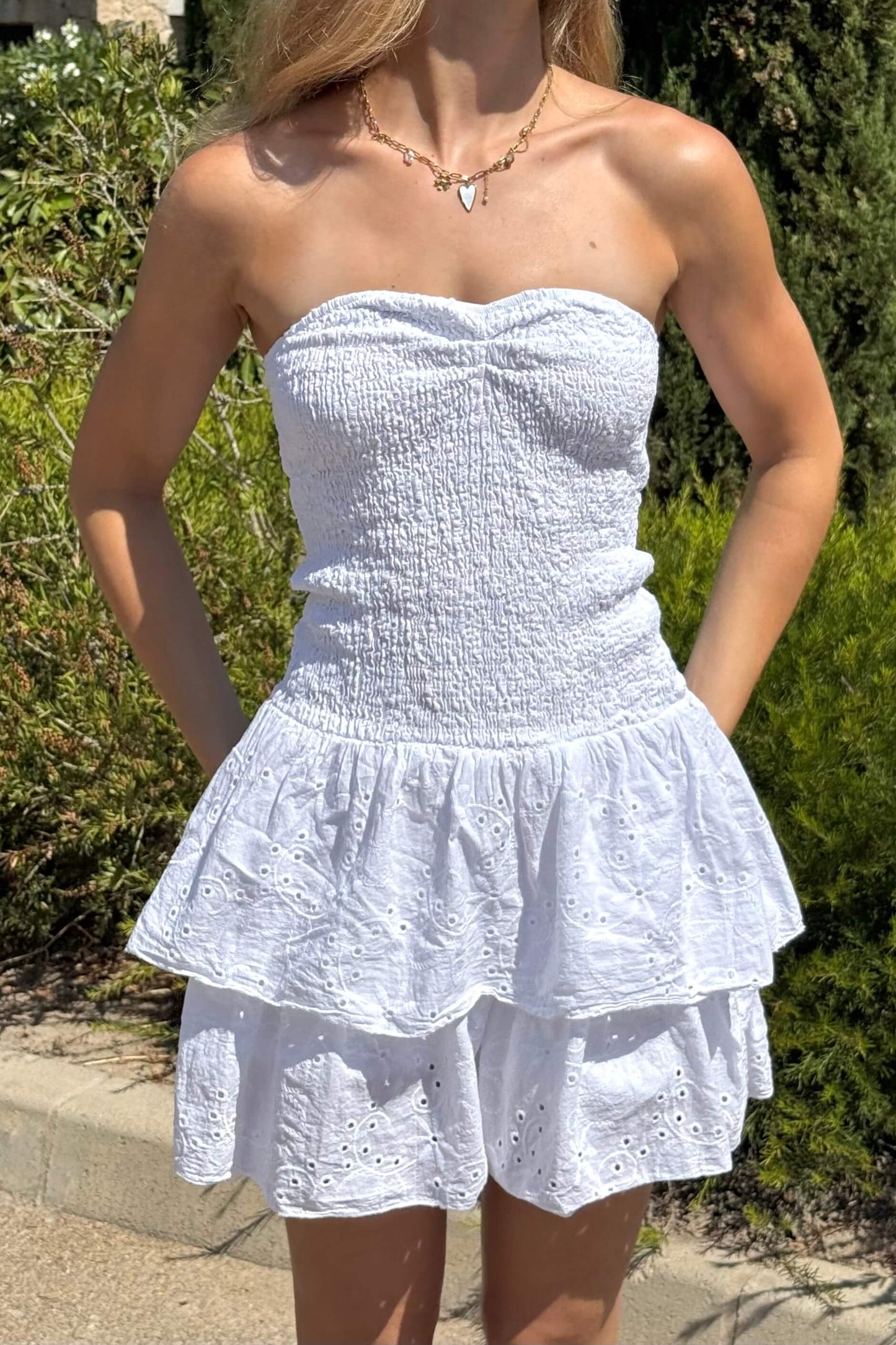 Robe bustier blanche femme en broderie anglaise portée en extérieur. Vue complète du modèle avec short intégré invisible. Coupe estivale, idéale pour l’été. Taille unique adaptée du S au L. Look frais et romantique pour plage, balade ou vacances.